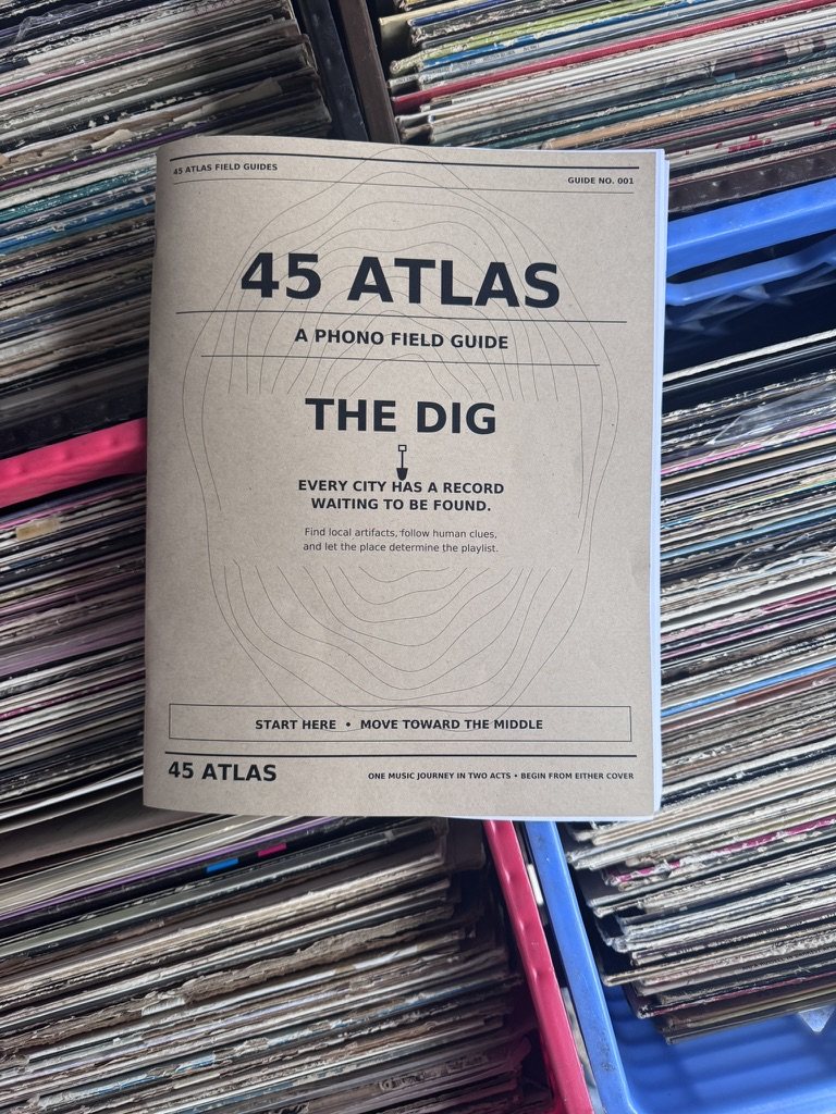 Tan-paper 45 Atlas: The Dig field guide resting across crates filled with vinyl records