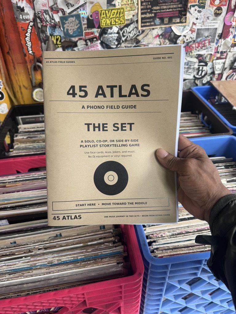 Barry Cole holding the tan-paper cover of 45 Atlas: The Set in front of vinyl crates and a sticker-covered record-store wall